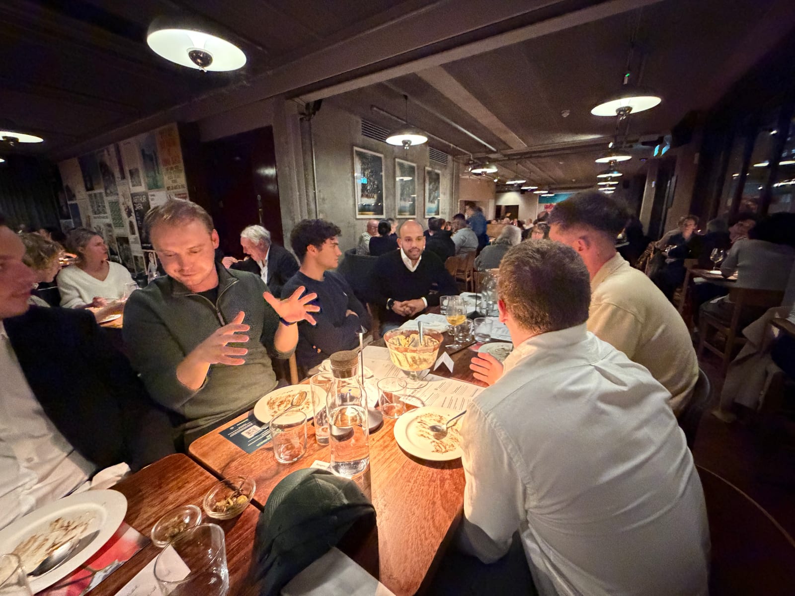 Guests mid-conversation around a long table in a dimly lit London restaurant, plates cleared and one person talking with their hands
